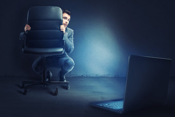 Afraid businessman hidden behind a chair looks his laptop