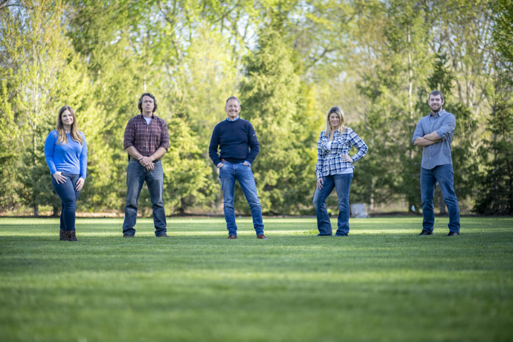 Portrait of five people standing on green grass in the sunlight.